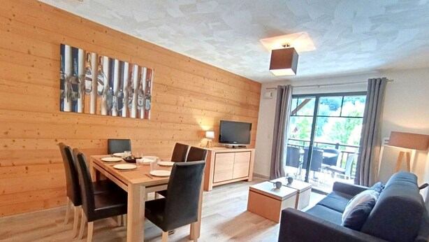 Una habitación moderna y bien iluminada con una mesa de comedor de madera, sillas negras, un sofá y un televisor sobre un mueble de madera.