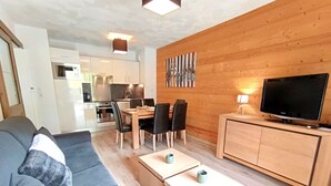 Una sala de estar moderna con un sofá, una mesa de comedor con sillas, un televisor sobre un mueble de madera y una cocina al fondo.
