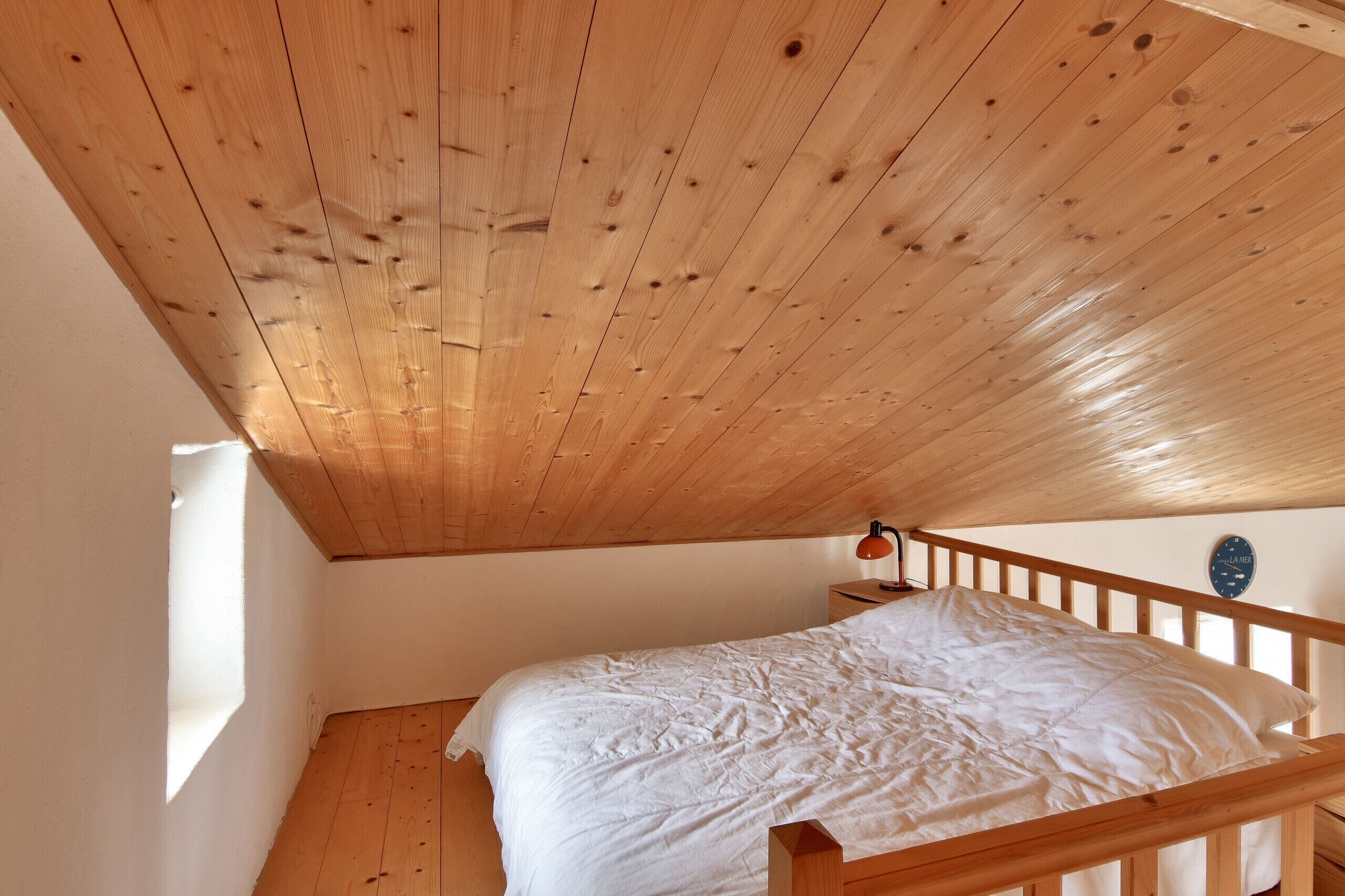 Un dormitorio con techo de madera, una cama con ropa blanca, una mesita de noche y un reloj de pared.