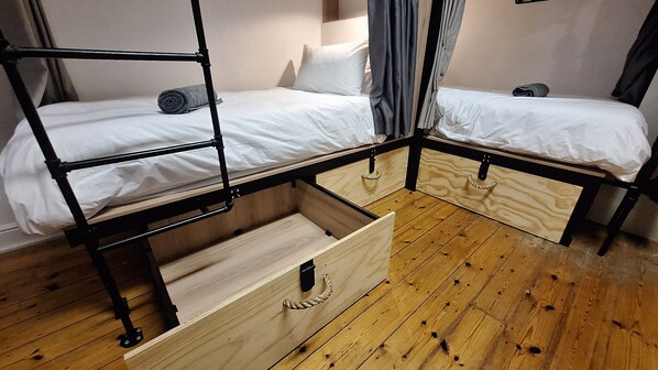 Habitación con literas, piso de madera, una cama individual con colchón blanco y una toalla enrollada, además de una litera inferior con estructura de madera.