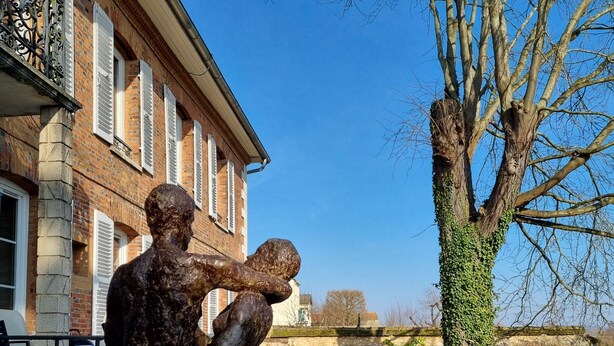 A bronze statue of two figures embracing, located in a courtyard with a brick building and a tree in the background. - Merlin Cottage (Montereau-Fault-Yonne)