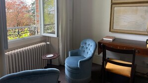 A room with a view of trees through a window, a radiator, a blue armchair, a wooden desk with a book, and a framed picture on the wall. - Merlin Cottage (Montereau-Fault-Yonne)