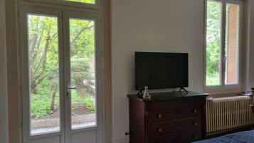 Ein Schlafzimmer mit Bett, Kommode, Fernseher und zwei Fenstern mit Blick auf Grünflächen.
