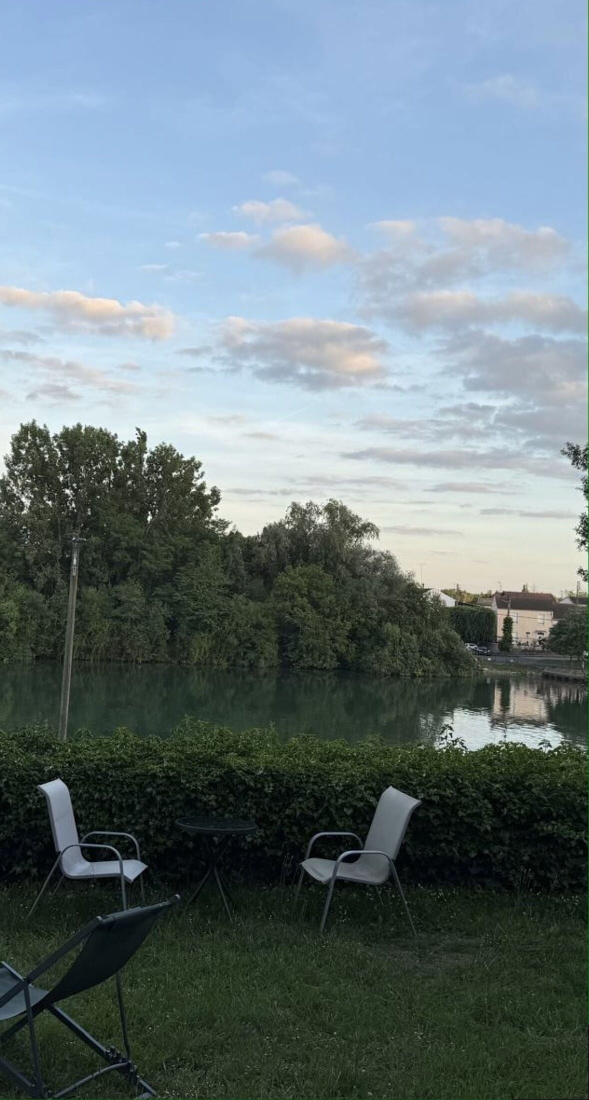 Vista a un lago con dos sillas y una mesa, rodeado de vegetación y un edificio al fondo.
