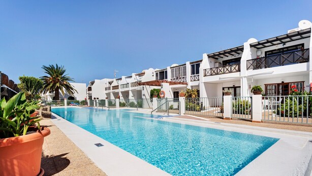 A swimming pool adjacent to a row of white buildings with balconies, surrounded by potted plants and a clear sky. - Apartment 'Romance' with Shared Pool, Private Terrace and Wi-Fi (Puerto del Carmen)
