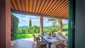 An outdoor dining area with a table set for four, surrounded by wicker chairs, under a wooden pergola with a view of a lush garden and distant hills. - Villa Belvedere on the Tuscan Hills (Toscana)
