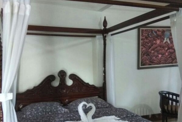 A four-poster bed with a heart-shaped towel arrangement, a framed floral painting on the wall, and a chair in the corner.