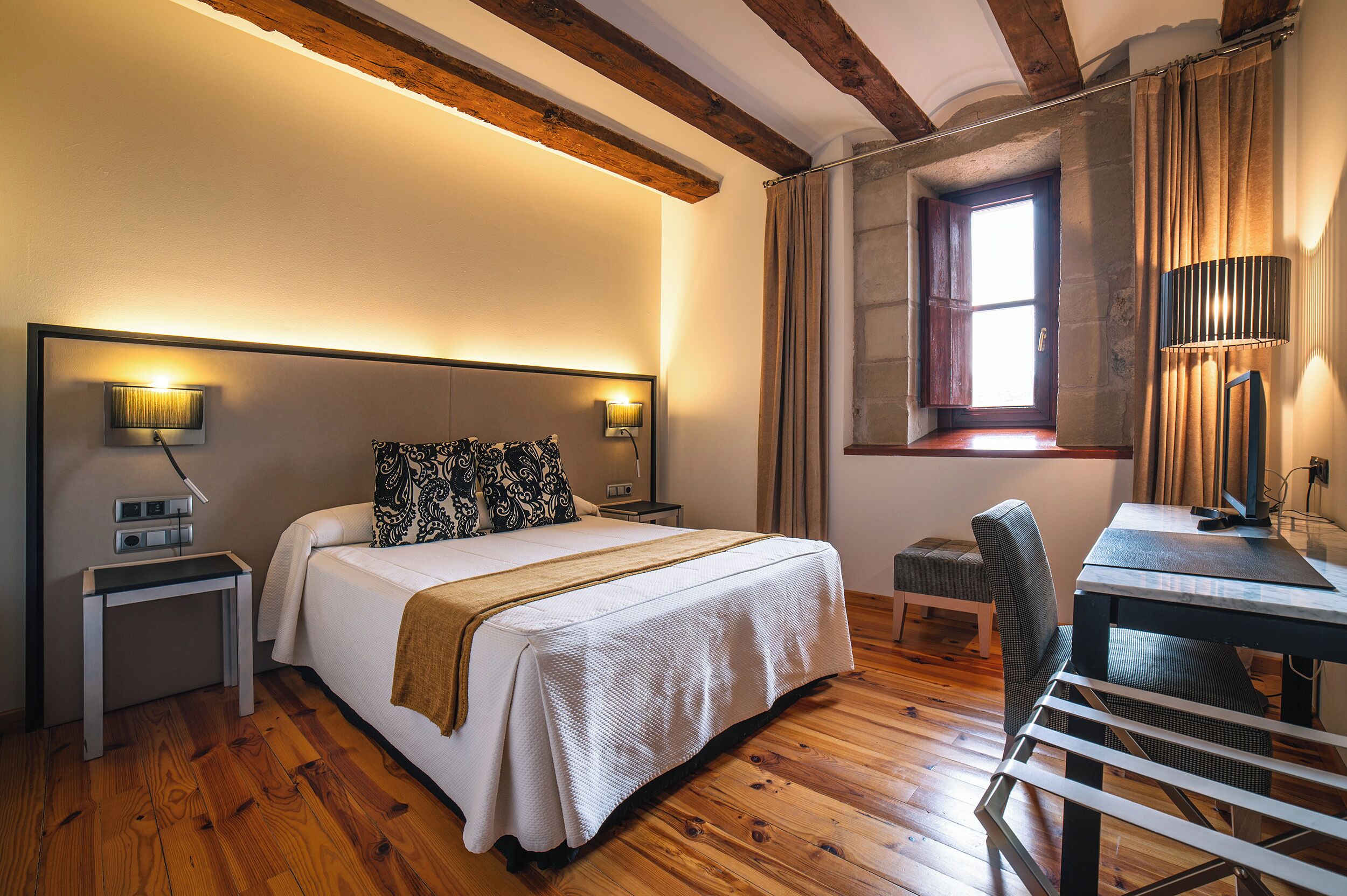 A hotel room with a wooden floor, a bed with a white bedspread and patterned pillows, a desk with a chair, and a window with curtains.