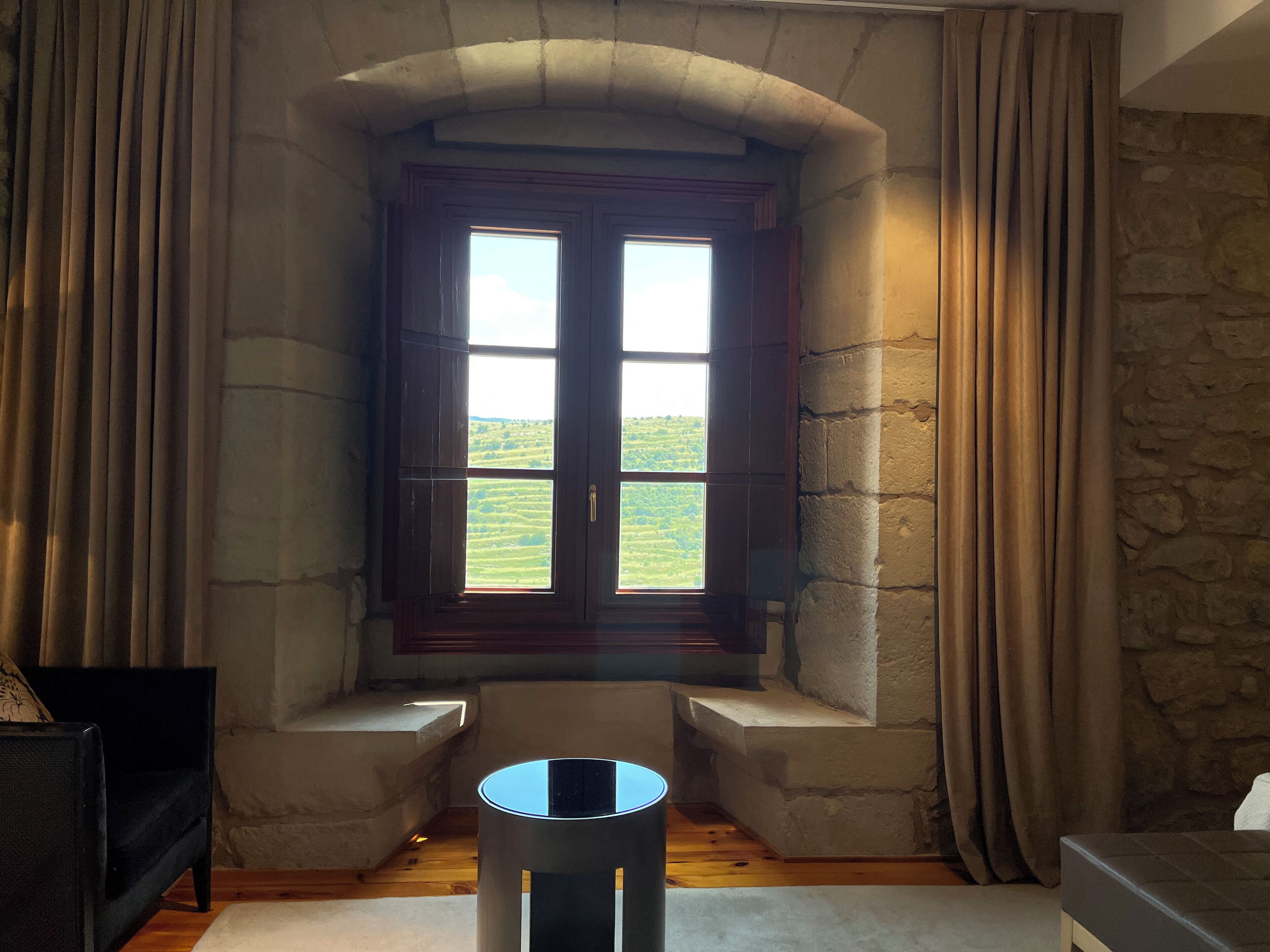 Una habitación con pared de piedra, una ventana con contraventanas de madera, una pequeña mesa redonda y un banco.
