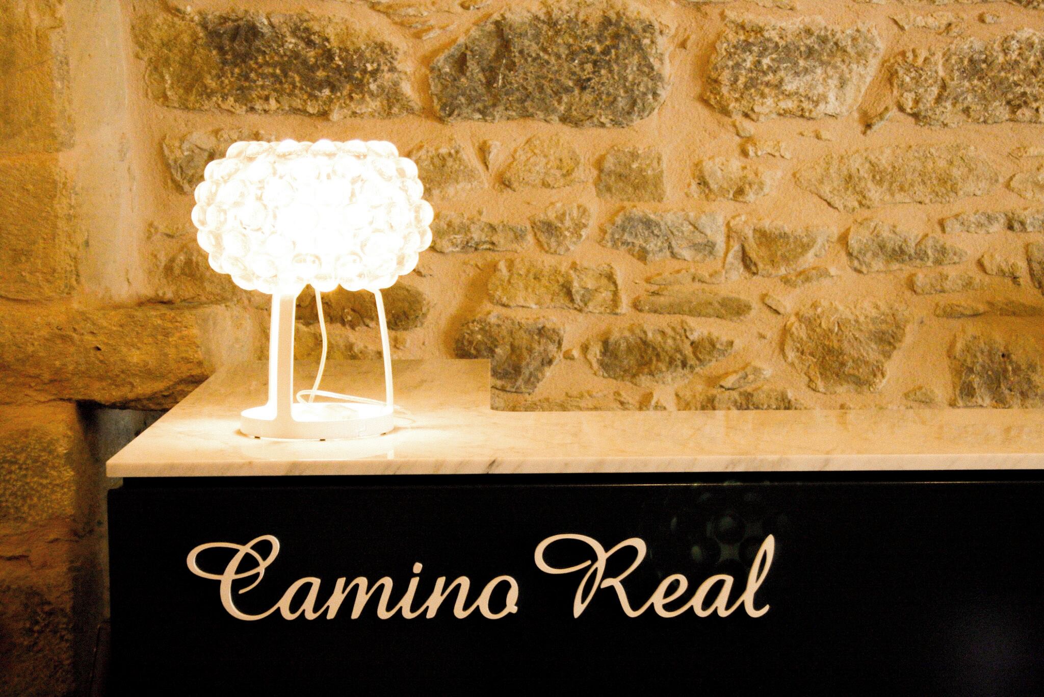 Un mur de pierre sur lequel est fixée une lampe blanche, le tout posé sur une surface noire portant l’inscription « Camino Real ».