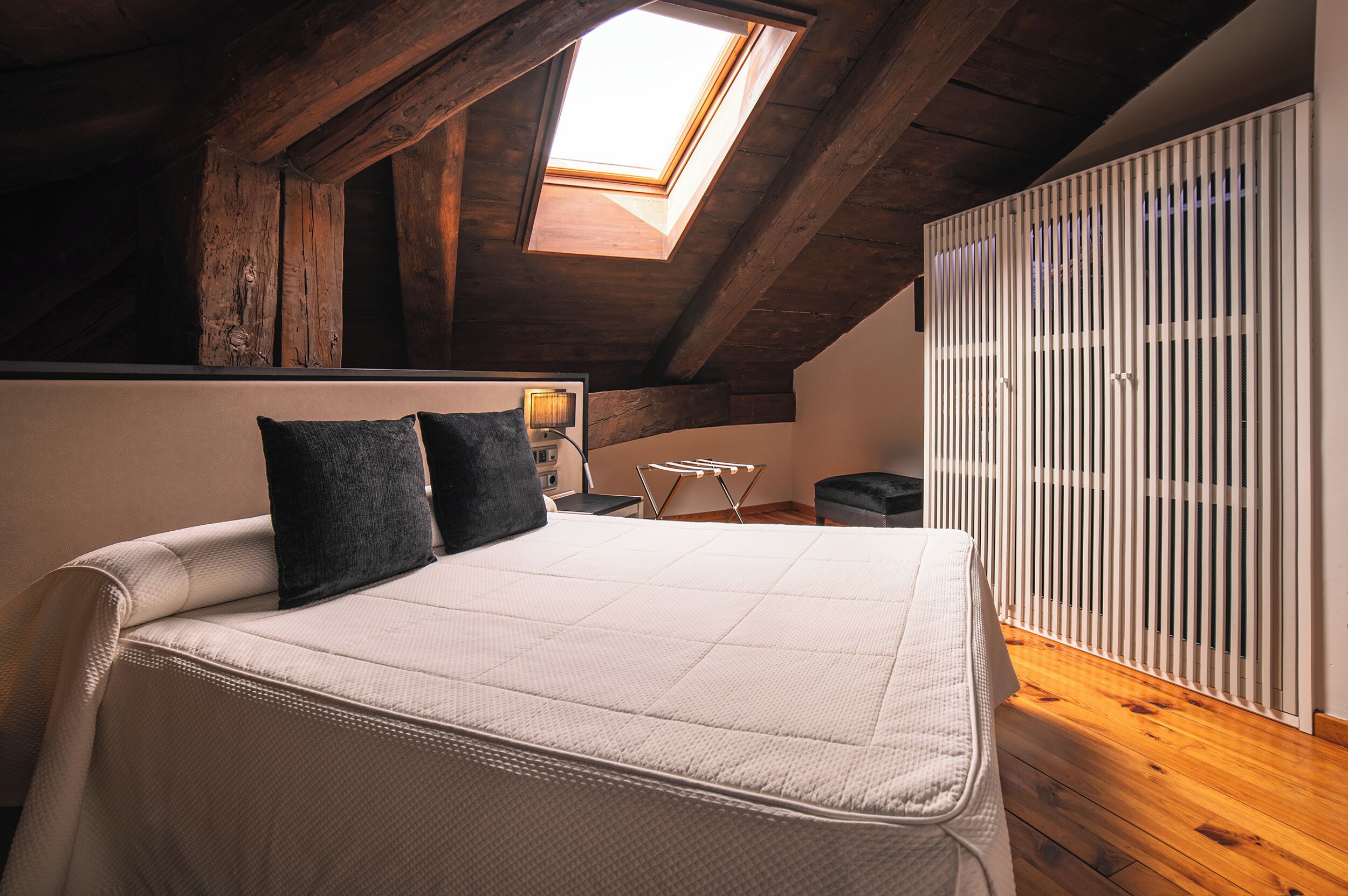 A bedroom with a sloped ceiling, a skylight, a bed with a white mattress and two pillows, a bedside table with a lamp, and a radiator.