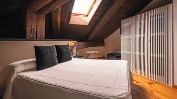 Un dormitorio con techo inclinado, tragaluz, cama con colchón blanco y dos almohadas, mesita de noche con lámpara y radiador.
