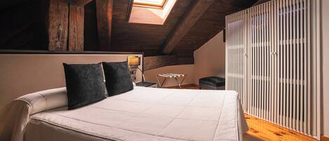 Un dormitorio con techo inclinado, tragaluz, cama con colchón blanco y dos almohadas, mesita de noche con lámpara y radiador.