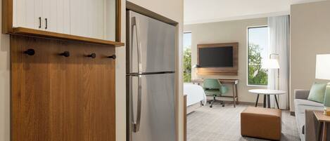 A hotel room with a wooden wardrobe, a stainless steel refrigerator, a bed with a green chair, a small table, a sofa, and a TV mounted on the wall.