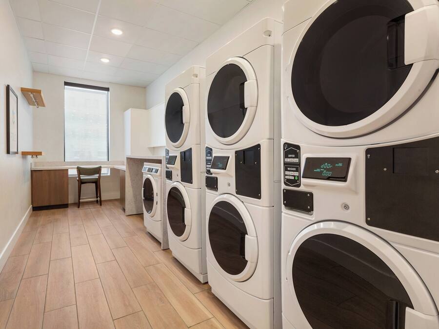 A laundry room with multiple washing machines and dryers, wooden flooring, and a window with blinds.