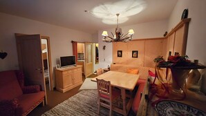 Una habitación acogedora y bien iluminada con una mesa de comedor, sillas, un televisor en un mueble de madera, un ventilador de techo y un jarrón decorativo con flores.