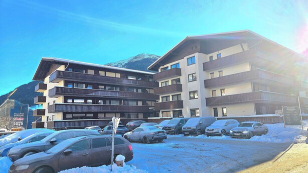 Un hotel de varios pisos con balcones, un estacionamiento con varios automóviles y un paisaje montañoso de fondo.