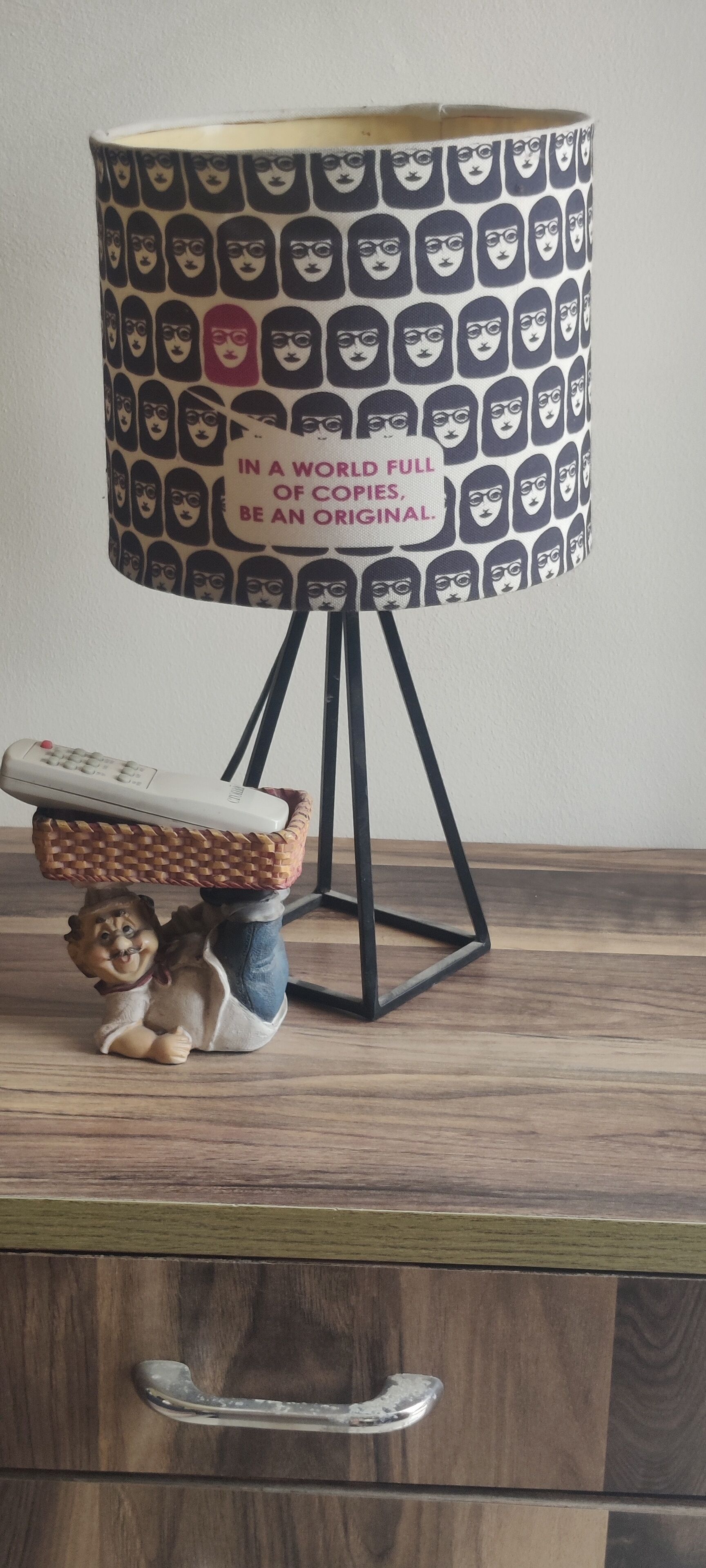 Une lampe avec un abat-jour à motifs et une citation, un panier contenant une télécommande et une figurine posée sur une surface en bois.