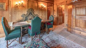 Uma sala de jantar tradicional em madeira, com mesa e cadeiras, arandelas ornamentadas e uma grande porta de madeira.