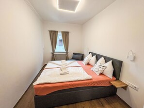 A neatly made bed with a dark headboard, a bedside table, and a view of the outside through a window with curtains.