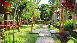 Un sendero en un jardín que lleva a un conjunto de columpios rodeado de exuberante follaje y plantas tropicales.