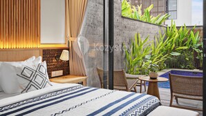 A modern hotel room with a large bed, a small table with a potted plant, wicker chairs, and a view of a swimming pool and greenery.
