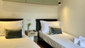 Habitación de hotel con dos camas individuales, cada una con ropa blanca y almohadas oscuras. Hay una mesita de noche pequeña con una lámpara y un jarrón con flores.