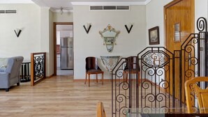 A living room with a wooden floor, a sofa, a dining table, and chairs. There is a staircase with a decorative railing.