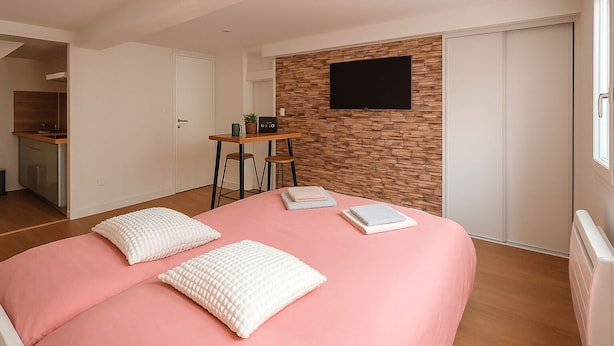 A bedroom with a pink bed, white pillows, a wooden wall panel, a TV mounted above the bed, and a small desk with a chair. - L'Escale Beauvaisienne - Hypercentre (Beauvais)