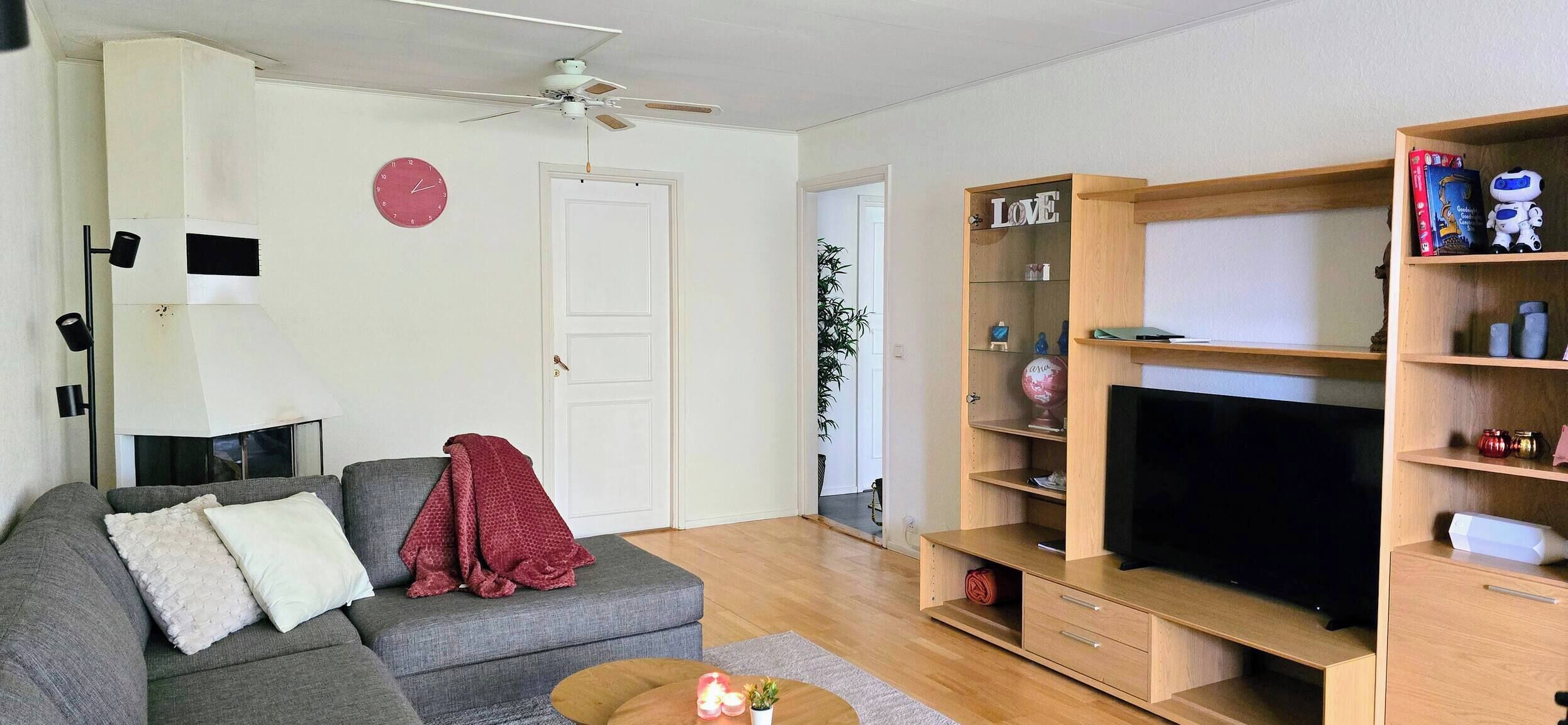 Una sala de estar con un sofá gris, un mueble para la televisión con estantes, un ventilador de techo y un reloj rojo en la pared.