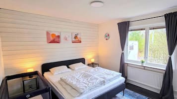 Quarto com cama, mesa de cabeceira, janela com cortinas e quadros nas paredes.