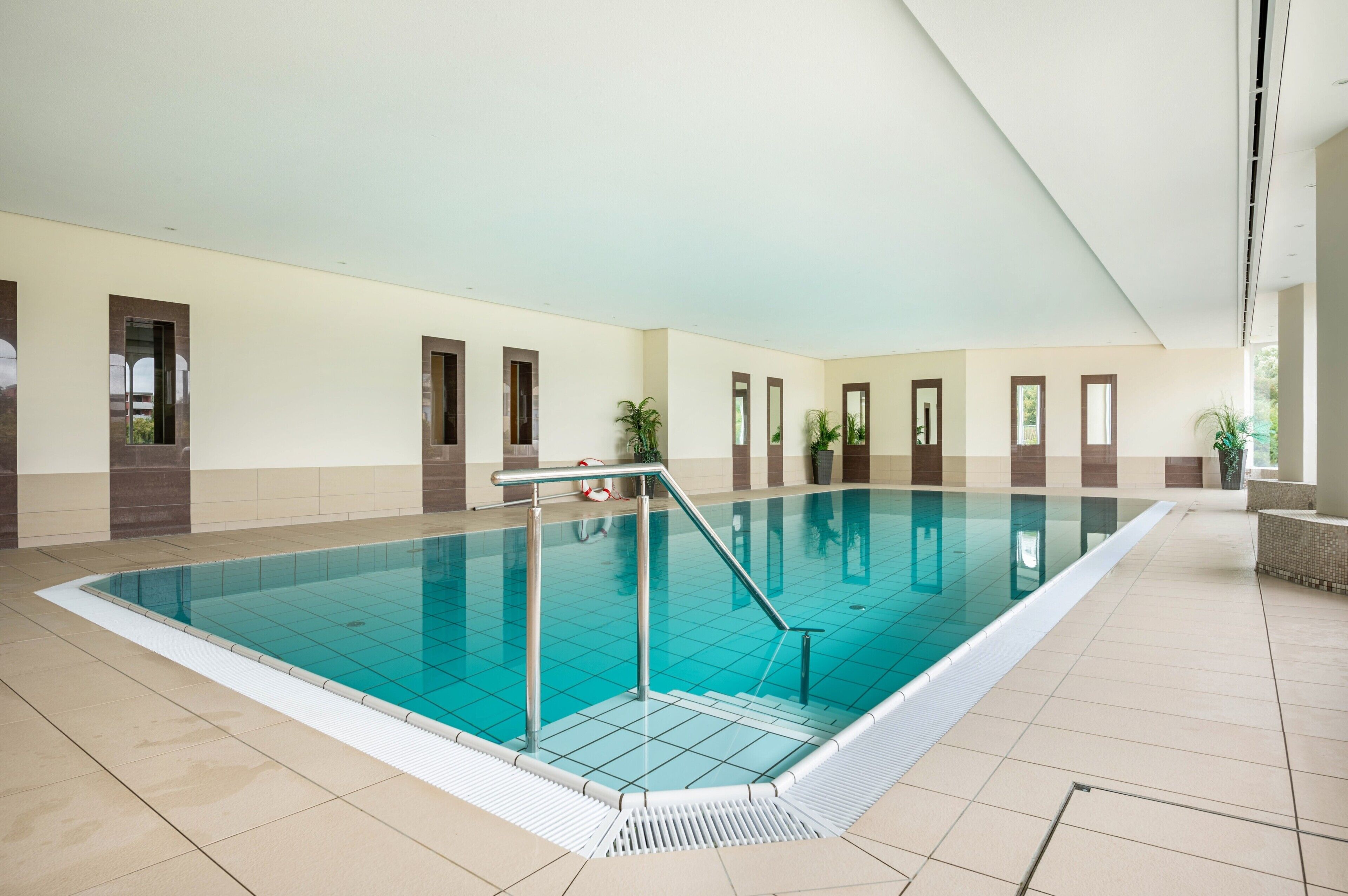 A large indoor swimming pool with a tiled floor, a metal pool ladder, and potted plants around the perimeter.