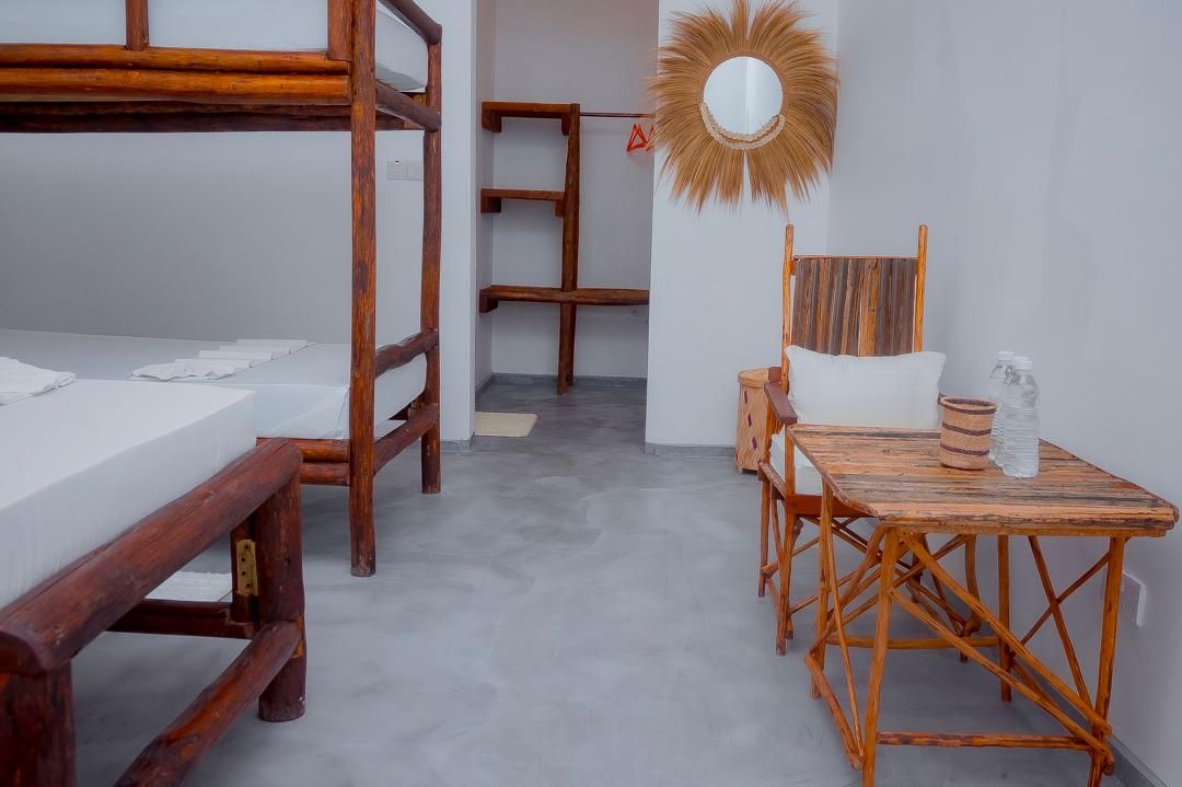 Una habitación con literas, una mesa de madera con sillas, un espejo y un estante pequeño.