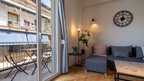 Ein modernes Wohnzimmer mit einer Couch, einem Couchtisch und einem Essbereich mit Stühlen und einem Tisch. Ein großes Fenster bietet Blick auf ein Gebäude, und es gibt einen Balkon.