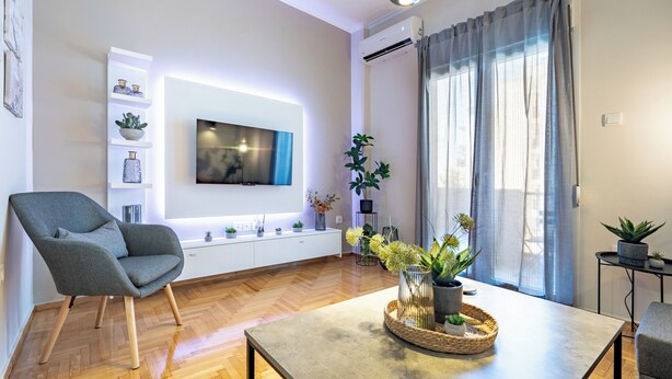 Ein modernes Wohnzimmer mit einem grauen Sessel, einem flachbildfernseher an der Wand, Holzboden und einem Esstisch mit einem gewebten Tablett.