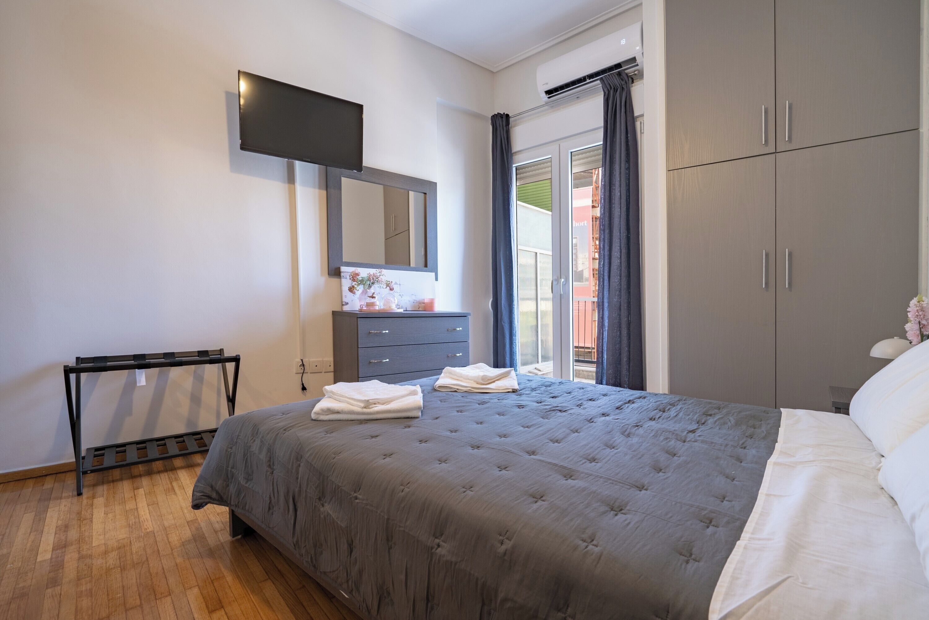 Une chambre à coucher comprenant un lit, une commode, un téléviseur fixé au mur et une fenêtre avec des rideaux.