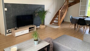 A modern living room with a flat-screen TV, wooden staircase, dining table, and a grey sofa.