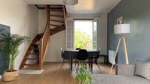 A modern living room with a wooden staircase, a dining area with a table and chairs, a grey sofa, and a floor lamp.