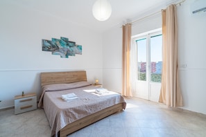 Un dormitorio con cama, mesita de noche, una obra de arte en la pared y vistas a través de una ventana con cortinas.
