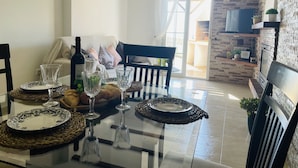 A dining area with a glass table set for four, featuring woven placemats and clear glassware. A bottle of wine and a bowl of fruit are on the table. The room has a stone wall and a television mounted on a brick wall. - White House with Sea Views by At Home Costa del Sol (Torrox Costa)