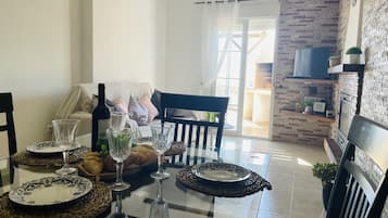 A dining area with a glass table set for four, featuring woven placemats and clear glassware. A bottle of wine and a bowl of fruit are on the table. The room has a stone wall and a television mounted on a brick wall.