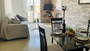 A modern living room with a sofa, a dining table set for two, and a kitchen area with a TV and brick wall. - White House with Sea Views by At Home Costa del Sol (Torrox Costa)