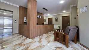A hotel reception area with a wooden reception desk, two clocks, a reception counter, a seating area with a chair, and a marble floor. - Ere's Palace (Tbilisi)