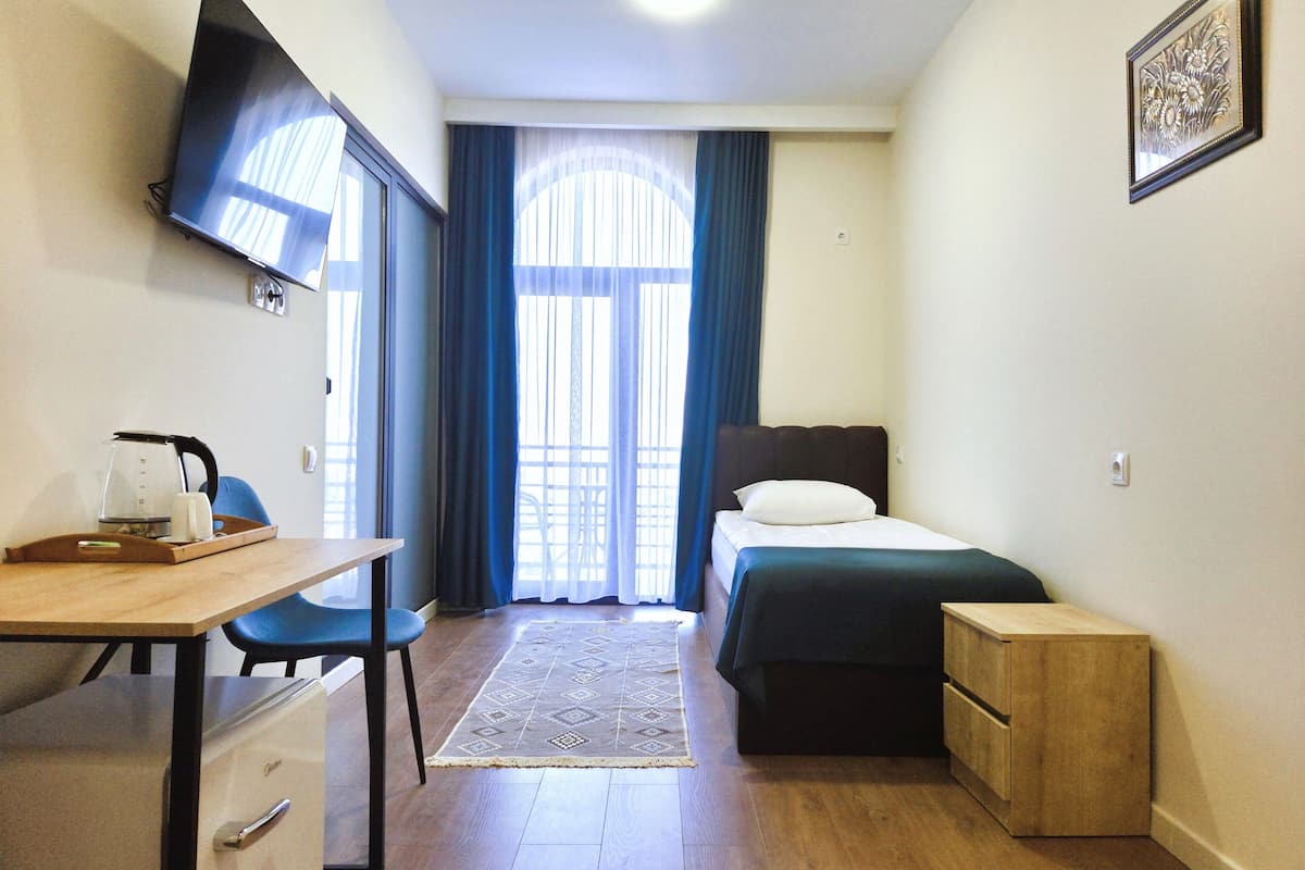 A compact hotel room with a bed, a desk with a chair, a small wooden side table, a television mounted on the wall, and a window with curtains.