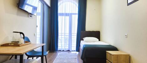 Une chambre d’hôtel compacte comprenant un lit, un bureau avec une chaise, une petite table d’appoint en bois, un téléviseur fixé au mur et une fenêtre avec des rideaux.