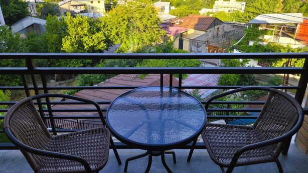 A balcony with two wicker chairs and a glass table, overlooking a cityscape with buildings and greenery. - Ere's Palace (Tbilisi)