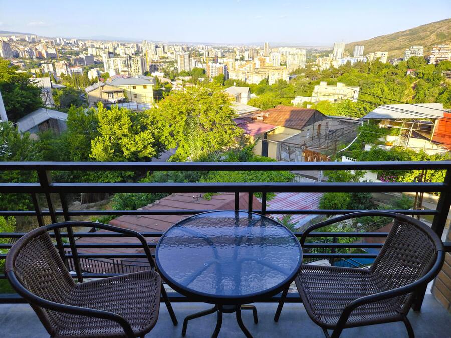 Un balcon avec deux fauteuils en osier et une table en verre, offrant une vue sur un paysage urbain avec des bâtiments et de la verdure.