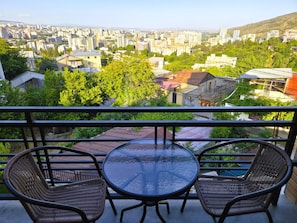 A balcony with two wicker chairs and a glass table, overlooking a cityscape with buildings and greenery. - Ere's Palace (Tbilisi)