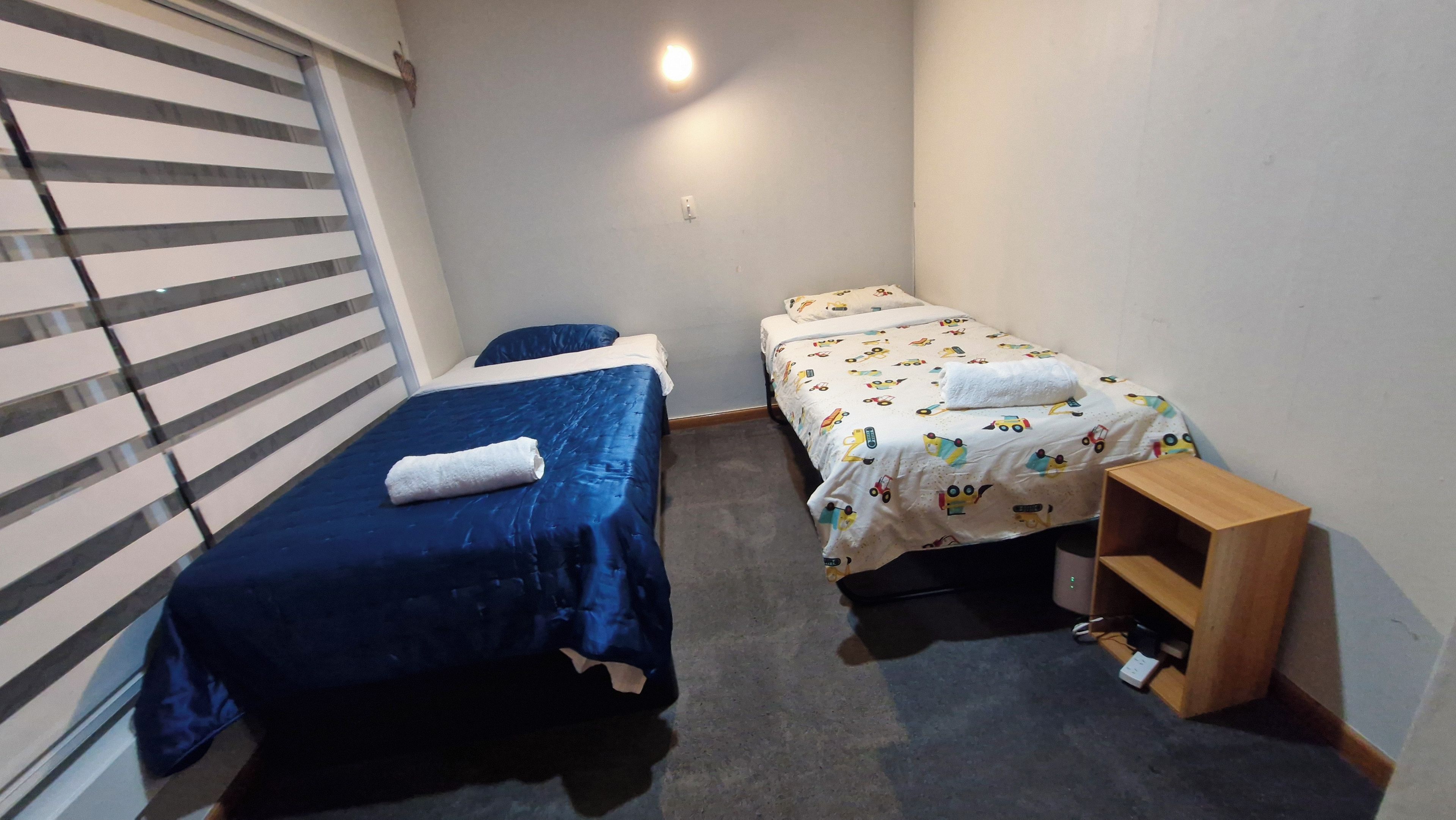 A small, single-room accommodation with two beds, a wooden nightstand, and a window with blinds.