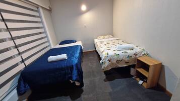 A small, single-room accommodation with two beds, a wooden nightstand, and a window with blinds.